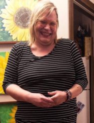 Burnunit a blonde femme person with glasses stands in front of a painting of sunflowers, clearly having a good time